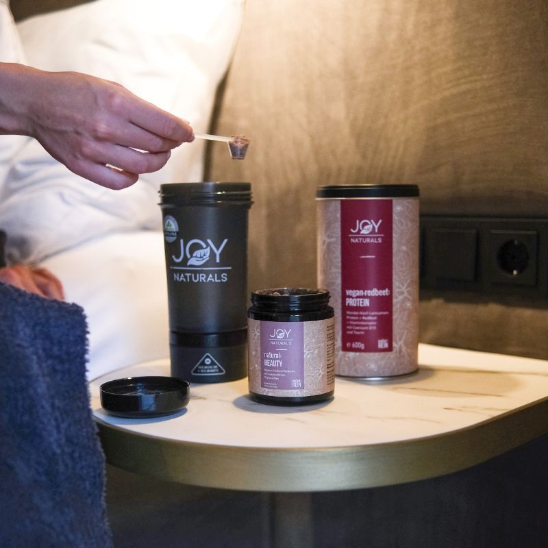 Tre contenitori su un tavolo. Barattolo: JOY NATURALS natural:BEAUTY Komplex. Barattolo: JOY NATURALS. Barattolo: proteine vegane di bacche rosse. Persona con misurino.