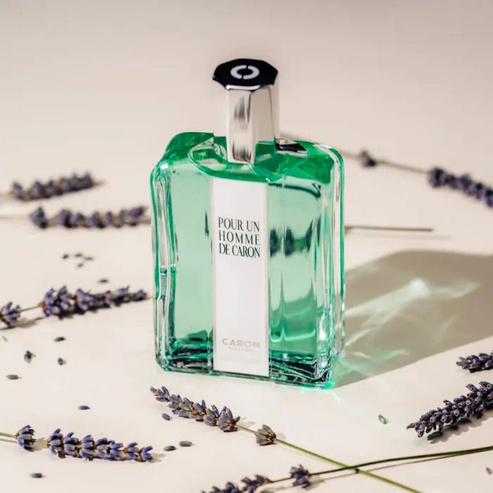 Flacone di Caron Pour Un Homme Eau De Toilette Spray. Flacone rettangolare, tappo argentato. Circondato da fiori di lavanda su sfondo chiaro.