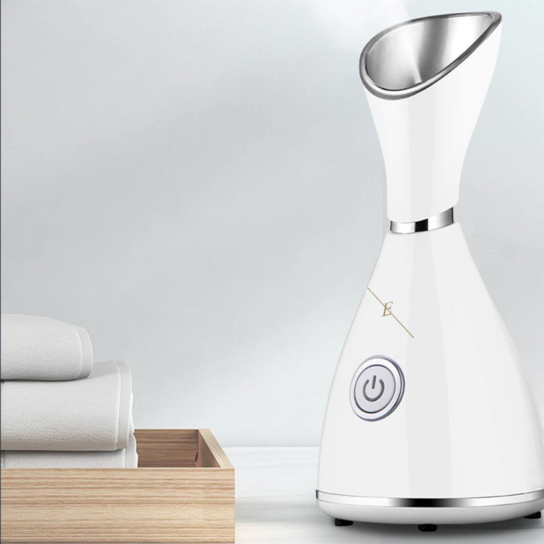 Vaporizzatore facciale bianco con testa a vapore argentata. Su un tavolo, accanto ad asciugamani e un vassoio di legno.