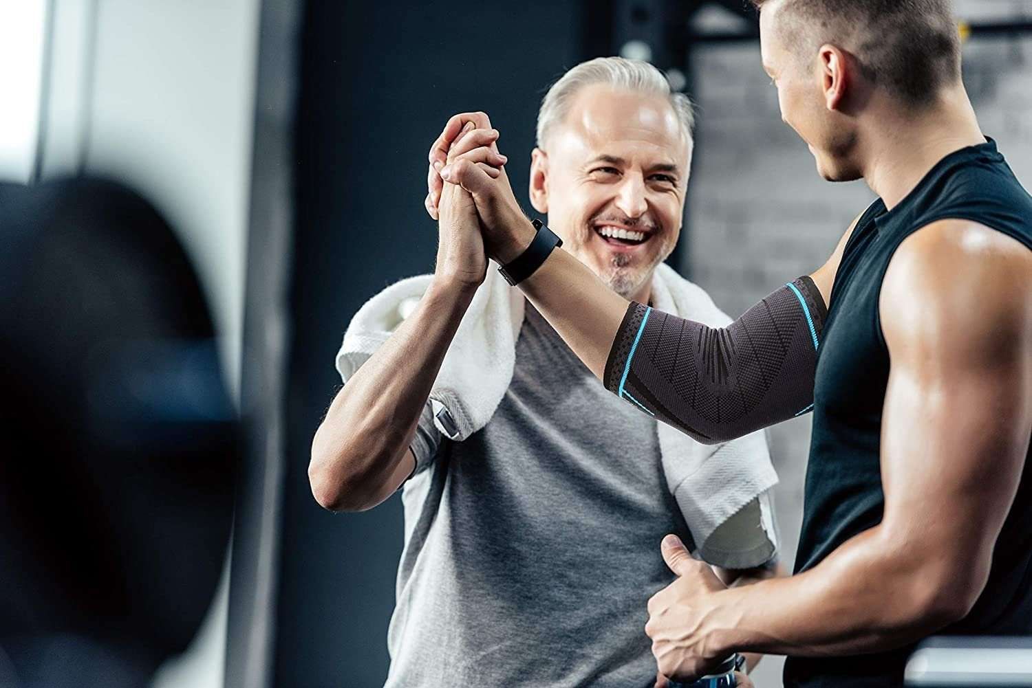 Due uomini in palestra. Uno indossa un tutore per gomito nero con dettagli blu. Si danno il cinque.