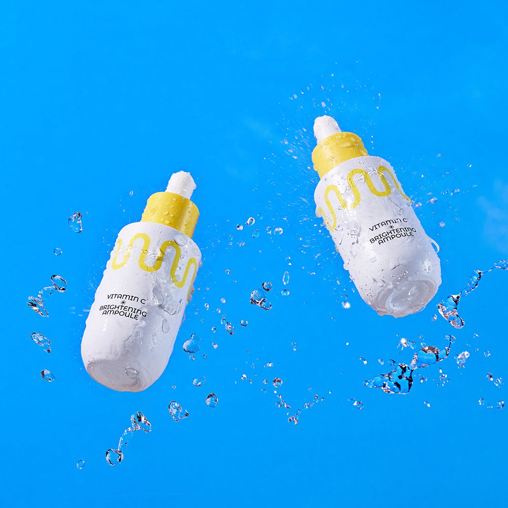 Due flaconi contagocce bianchi con tappi gialli, circondati dall'acqua. Contiene Vitamin C Brightening Ampoule.