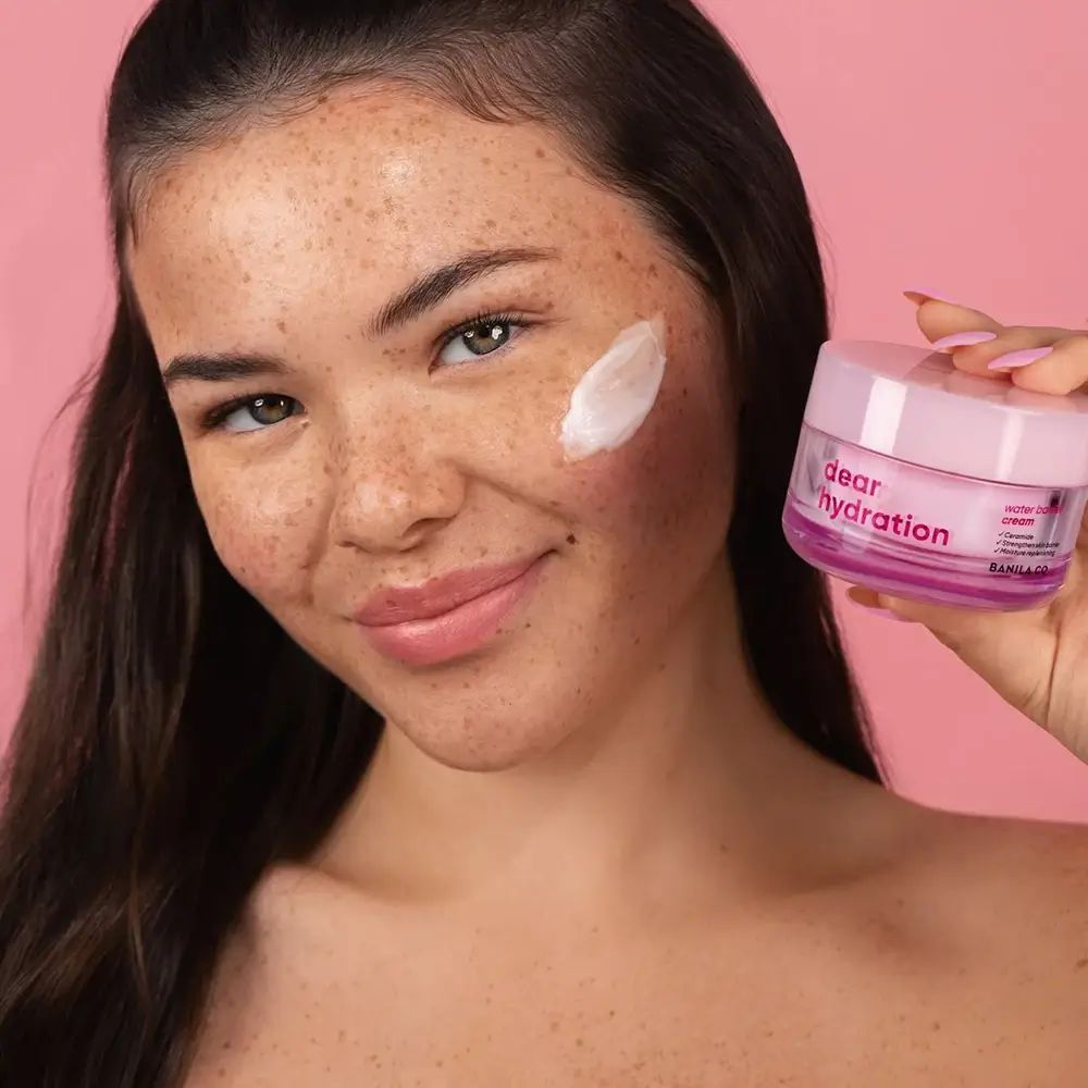 Donna con crema sulla guancia, tiene un vasetto. Sul vasetto c'è scritto "dear hydration" e "BANILA CO".