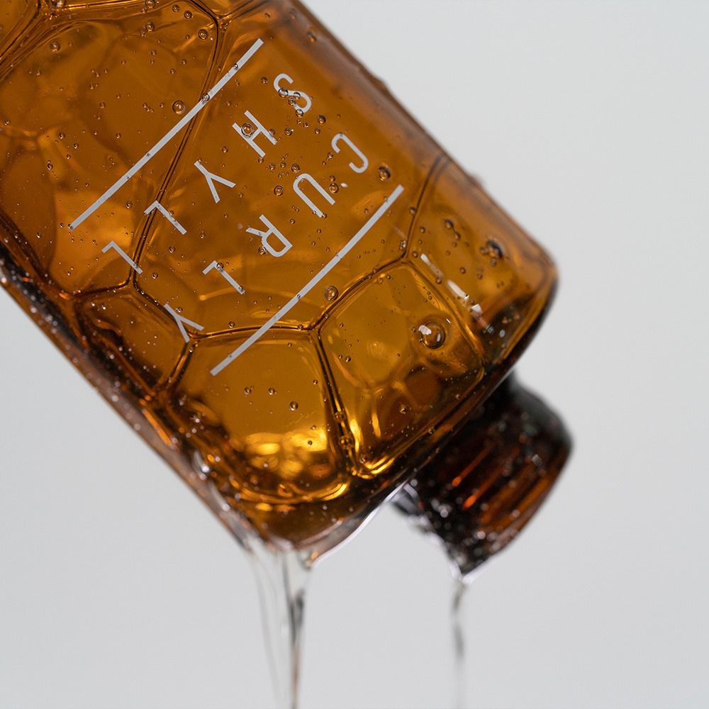 Flacone di shampoo marrone, da cui fuoriesce liquido. Testo: CURLY SHYLL.