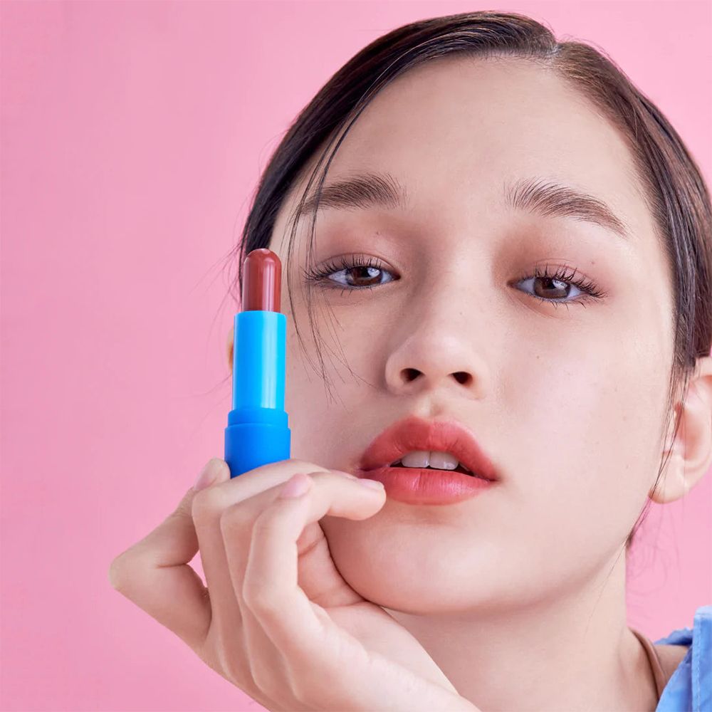 Persona tiene balsamo per labbra. Il balsamo ha un involucro blu e una punta rossa. Sfondo rosa.