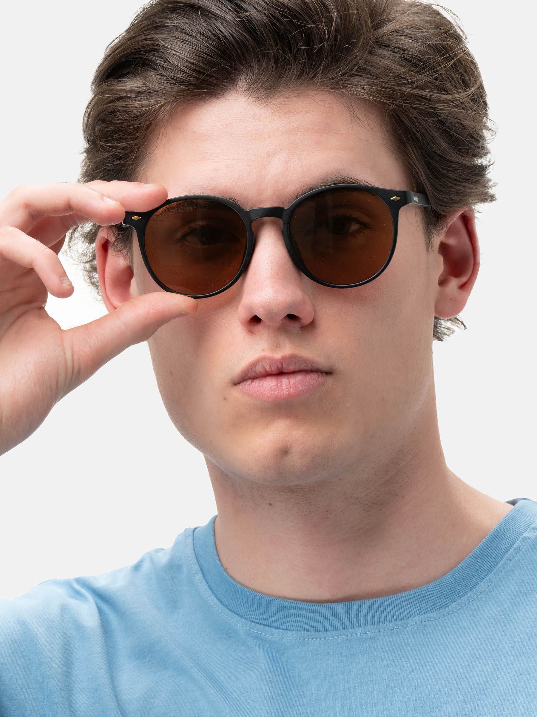 Uomo con occhiali da sole. Montatura nera, lenti marroni. Marchio 'smooder' sulla stanghetta. Sfondo blu.