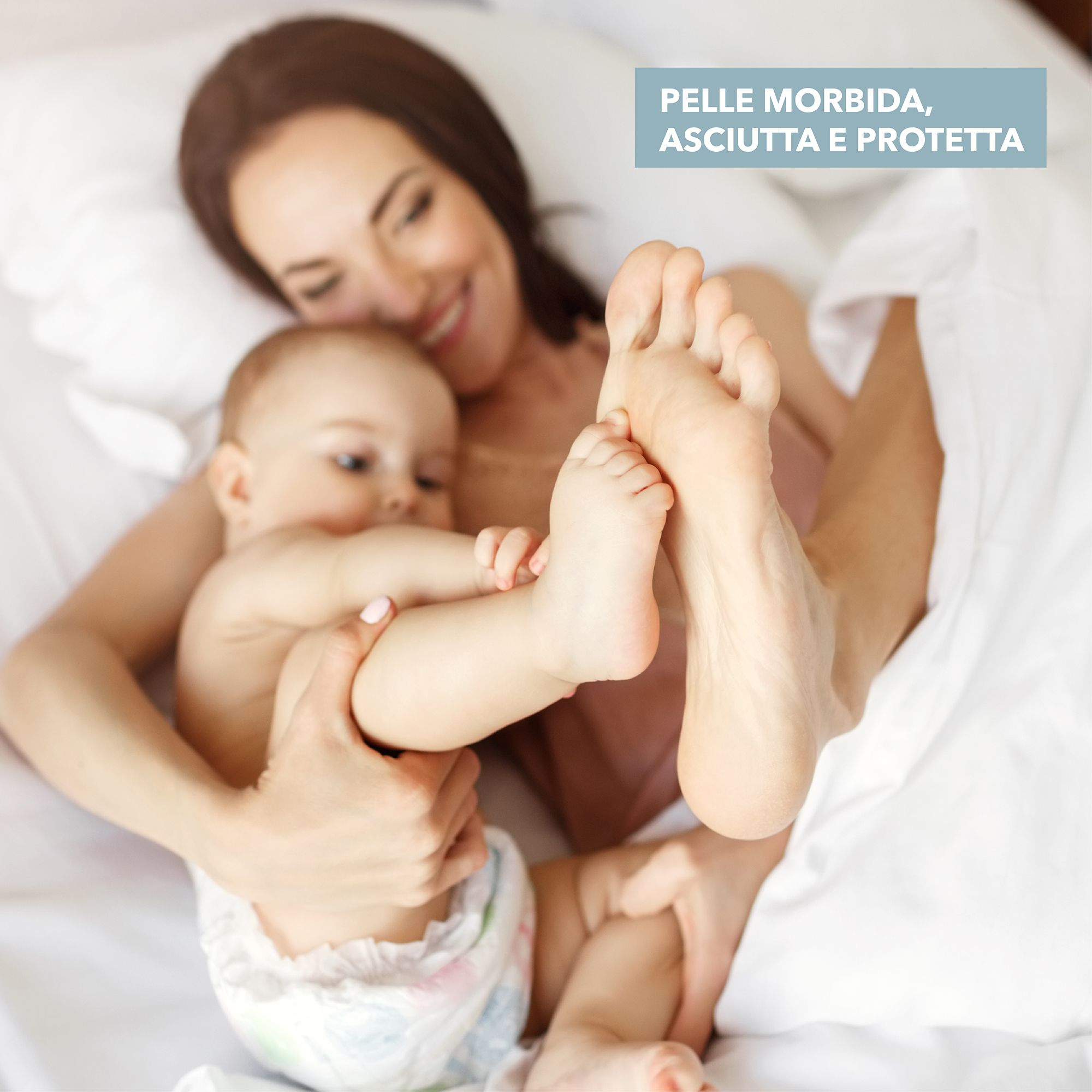 Mamma e bambino a letto. Piedi del bambino in primo piano. Testo: Pelle morbida, asciutta e protetta. Il bambino indossa un pannolino.