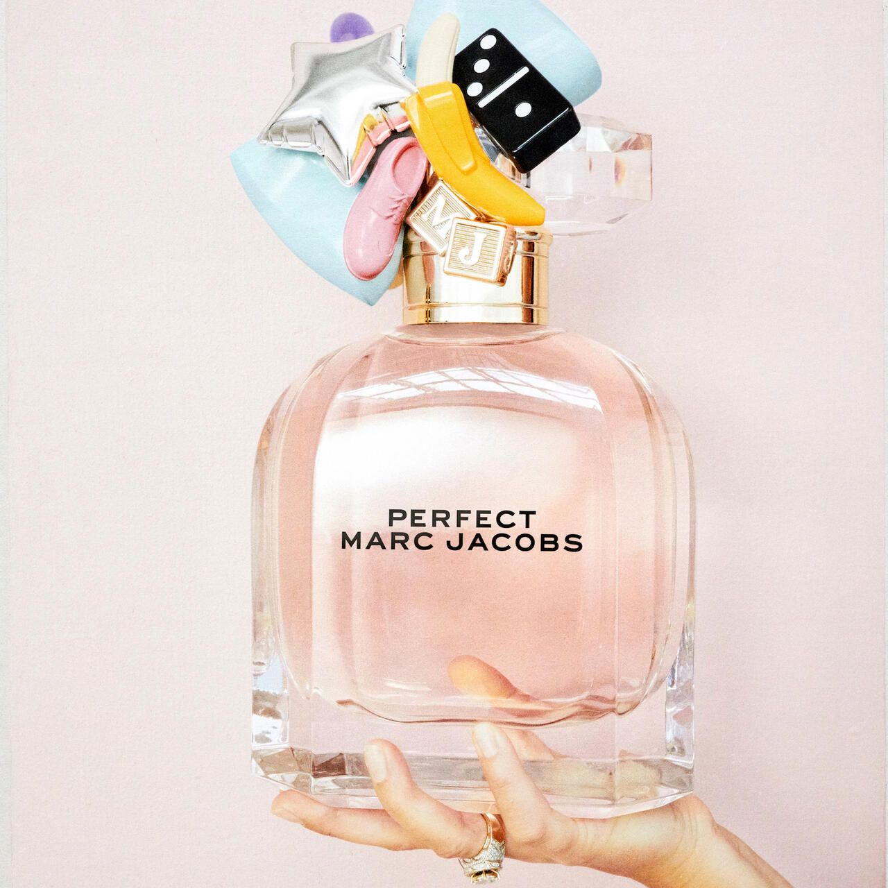 Flacone di profumo con tappo decorativo. Scritta: PERFECT MARC JACOBS. Flacone tenuto da una mano. Anello al dito.