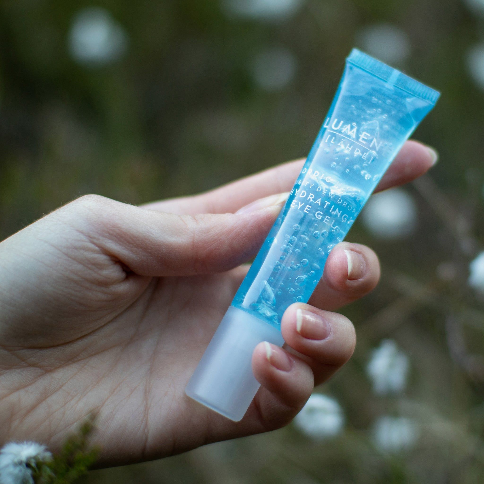 Mano che tiene un tubo blu. Scritta: Lumene [LAHDE] Nordic Hydra Purity Dew Drops Hydrating Eye Gel. Sfondo: fiori sfocati.