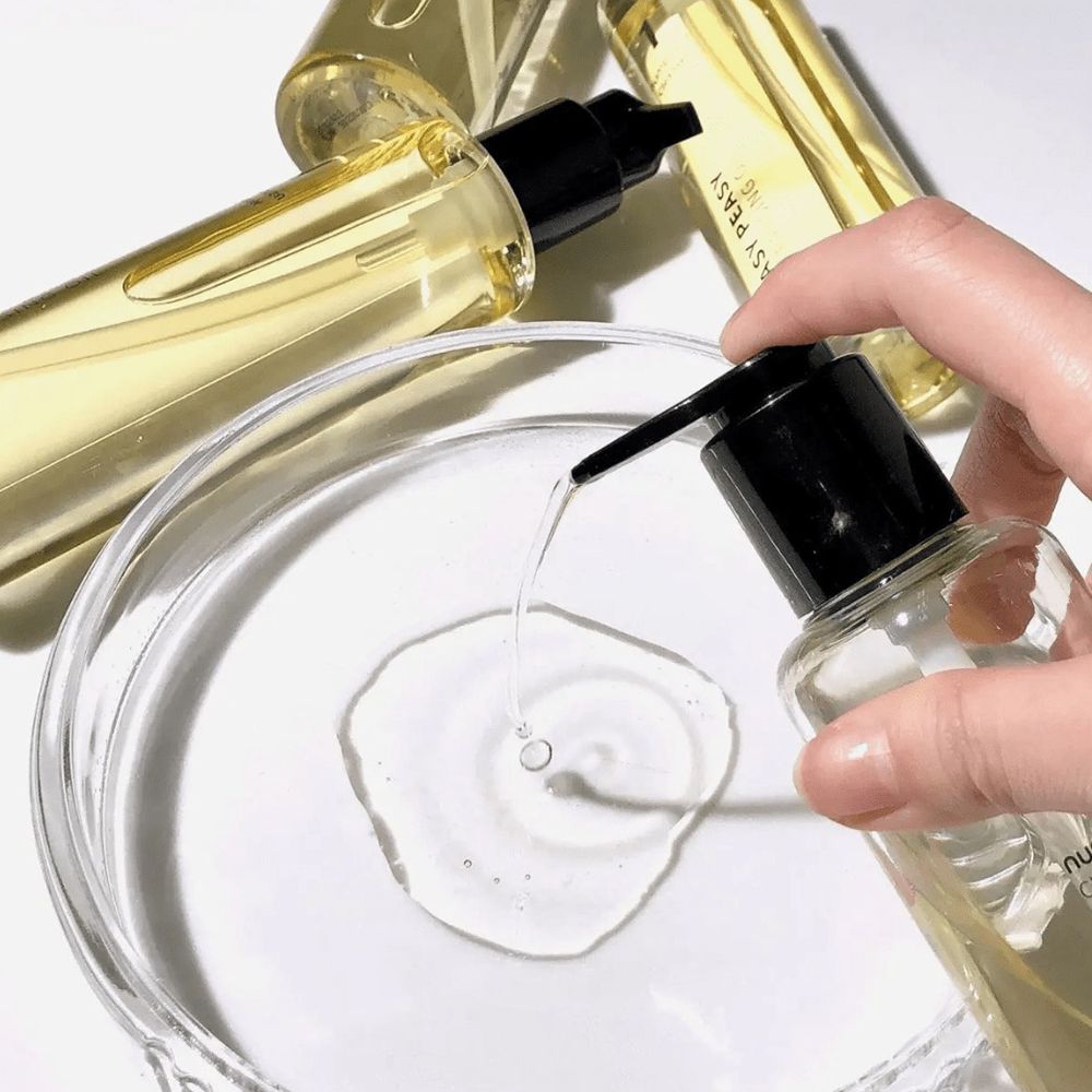 Una mano tiene un flacone di olio detergente sopra una ciotola di vetro. L'olio viene pompato. Diversi flaconi sullo sfondo.