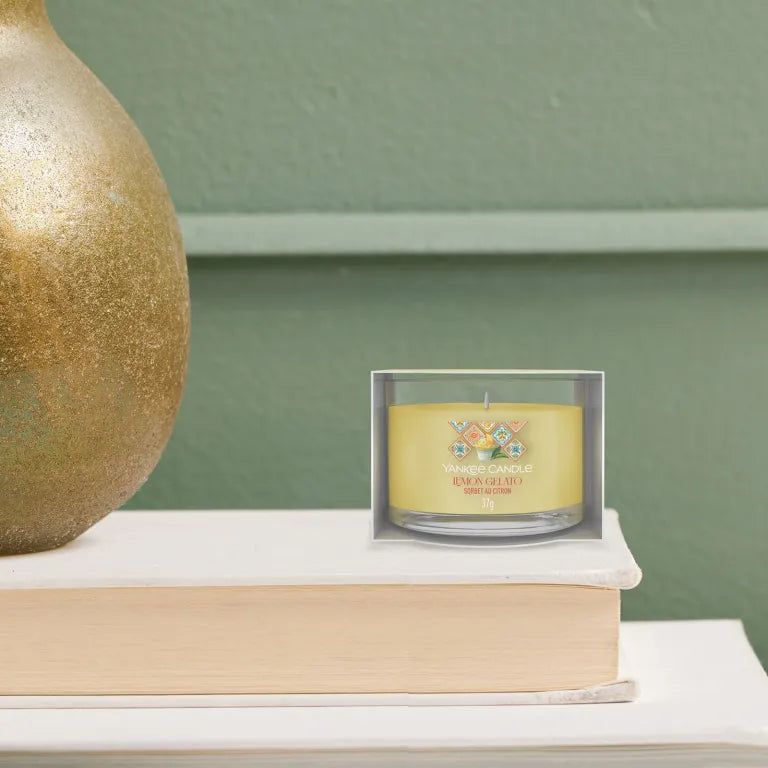 Candela gialla in vetro nella confezione. Scritta: Lemon Gelato. Posizionata su un libro. Sfondo: parete verde.