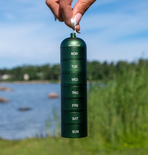 Portapillole verde a 7 scomparti, tenuto davanti a uno sfondo naturale con acqua ed erba.