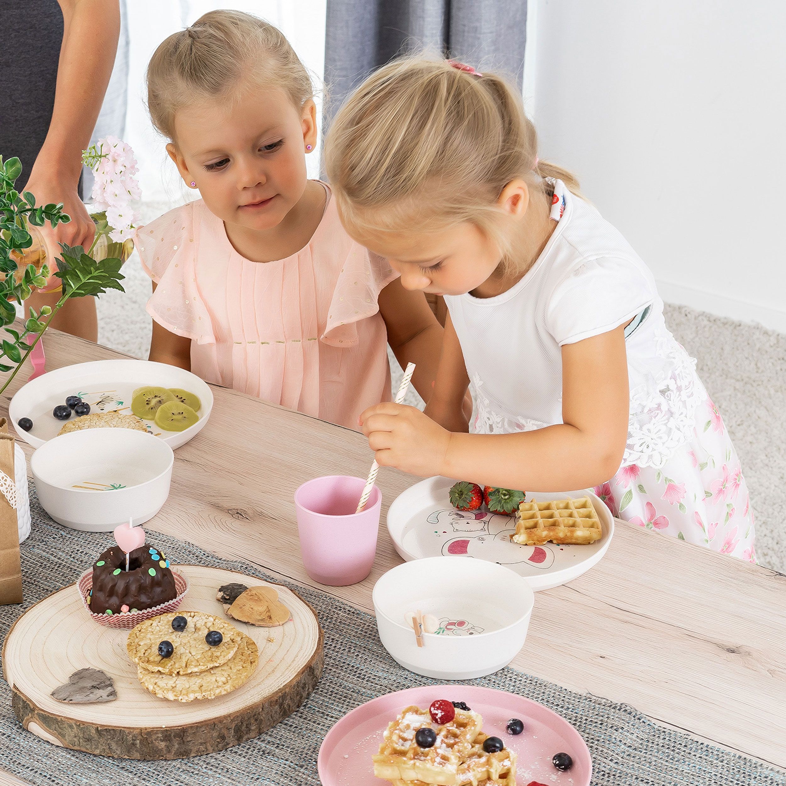 Due ragazze sedute a tavola con stoviglie per bambini. Piatti con waffle, bacche e torte. Tazze rosa e bianche.