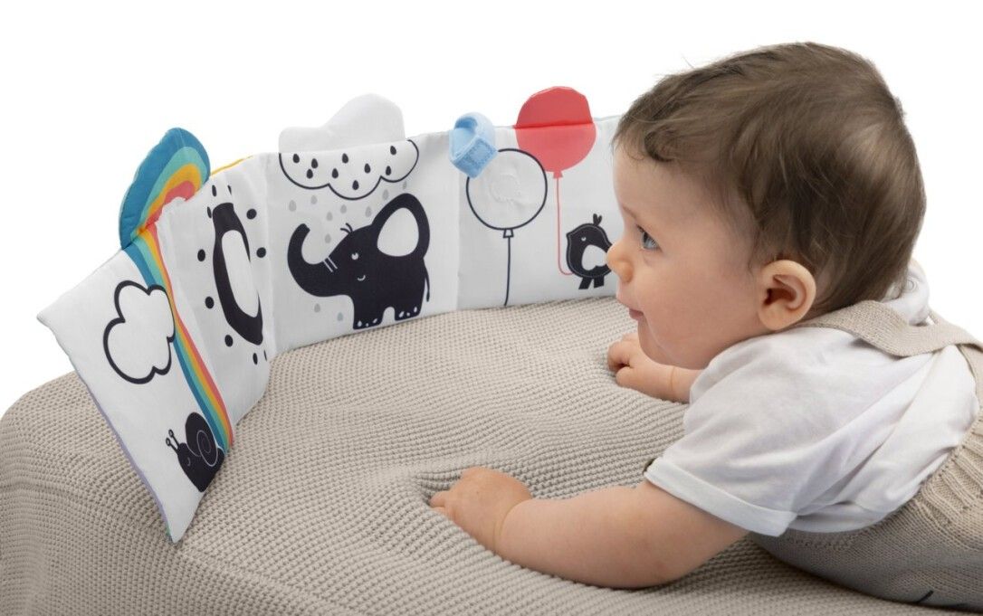 Bambino sdraiato su una coperta guarda un libro attività morbido. Libro con motivi animali e naturali. Illustrazioni in bianco e nero.