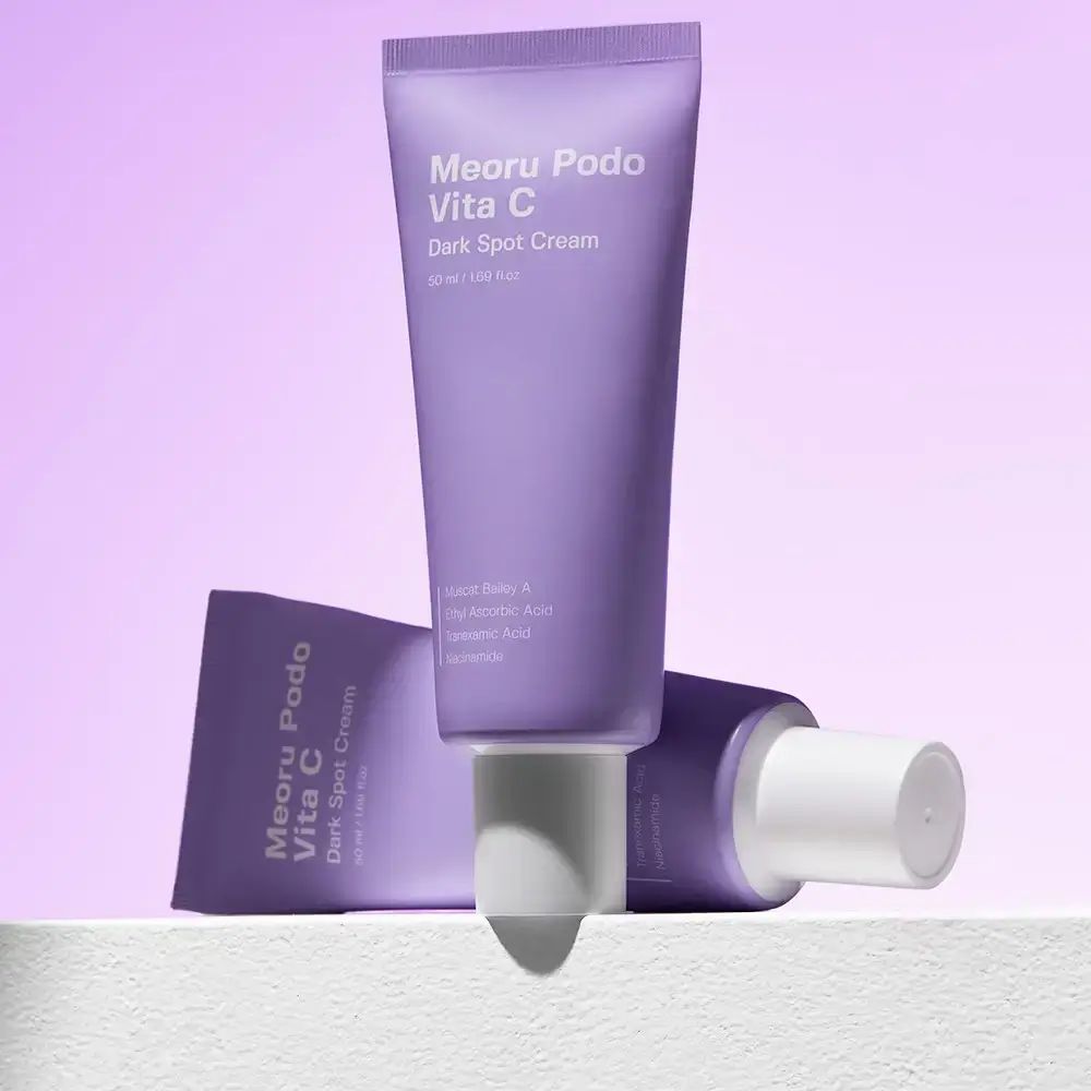 Due tubi viola 'Meoru Podo Vita C Dark Spot Cream'. Uno in piedi, l'altro sdraiato. Sfondo bianco.