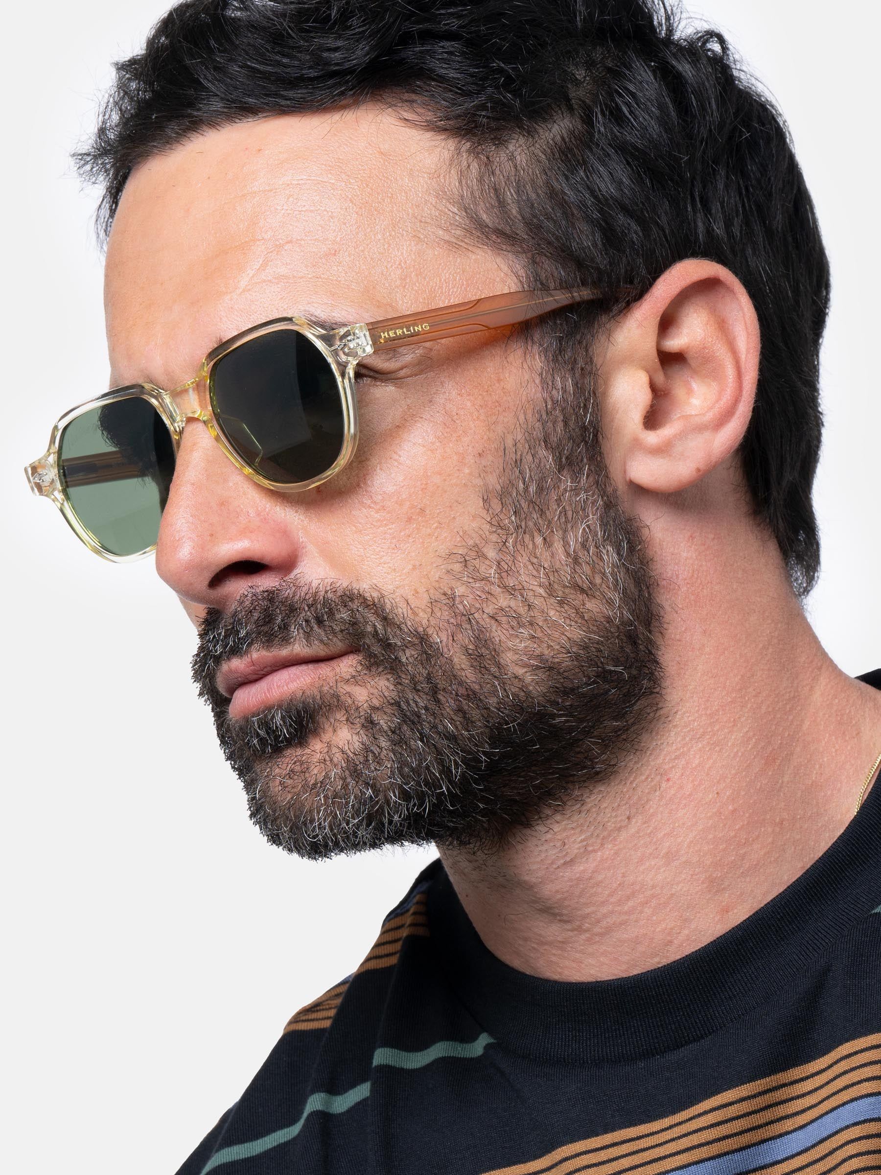 Uomo con occhiali da sole Herling con montatura giallo chiaro e lenti verdi. Aste marroni. Logo Herling visibile.