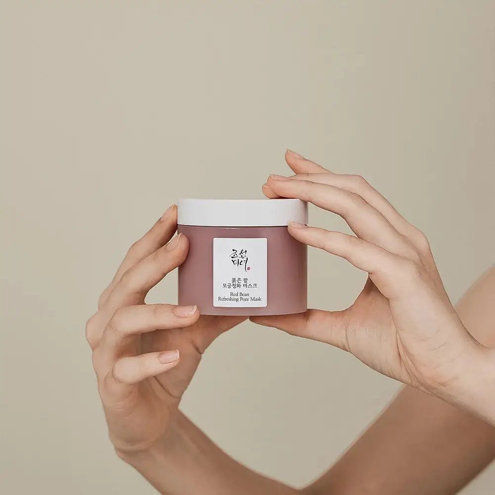 Mani che tengono un vaso rotondo con coperchio bianco. Scritte: caratteri coreani, Red Bean Refreshing Pore Mask. Contenuto rosa.