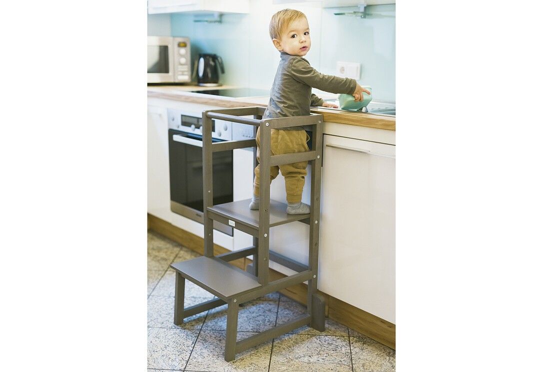 Bambino in piedi su una torre di apprendimento grigia in cucina. Il bambino afferra un oggetto.