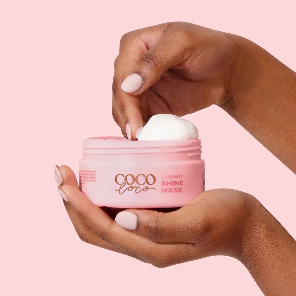 Mani che tengono un barattolo rosa di maschera per capelli al cocco. Un dito preleva la crema. Scritta: Coco Loco Shine Mask.