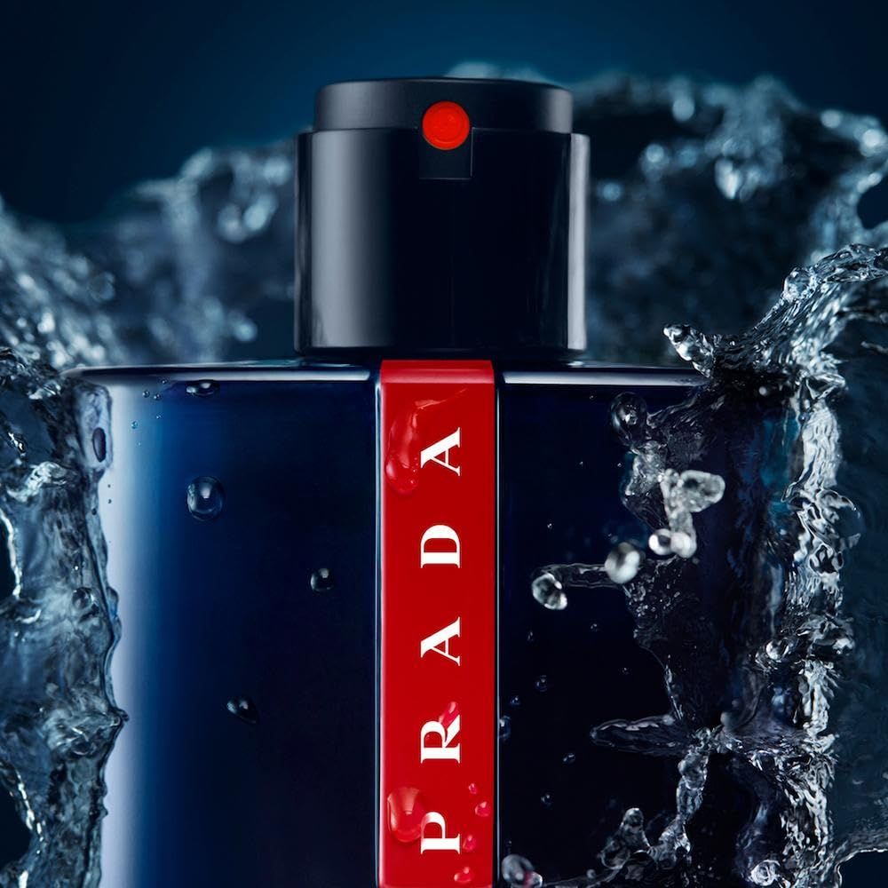 Flacone blu scuro, striscia rossa Prada. Erogatore nero. Spruzzi d'acqua. Primo piano.
