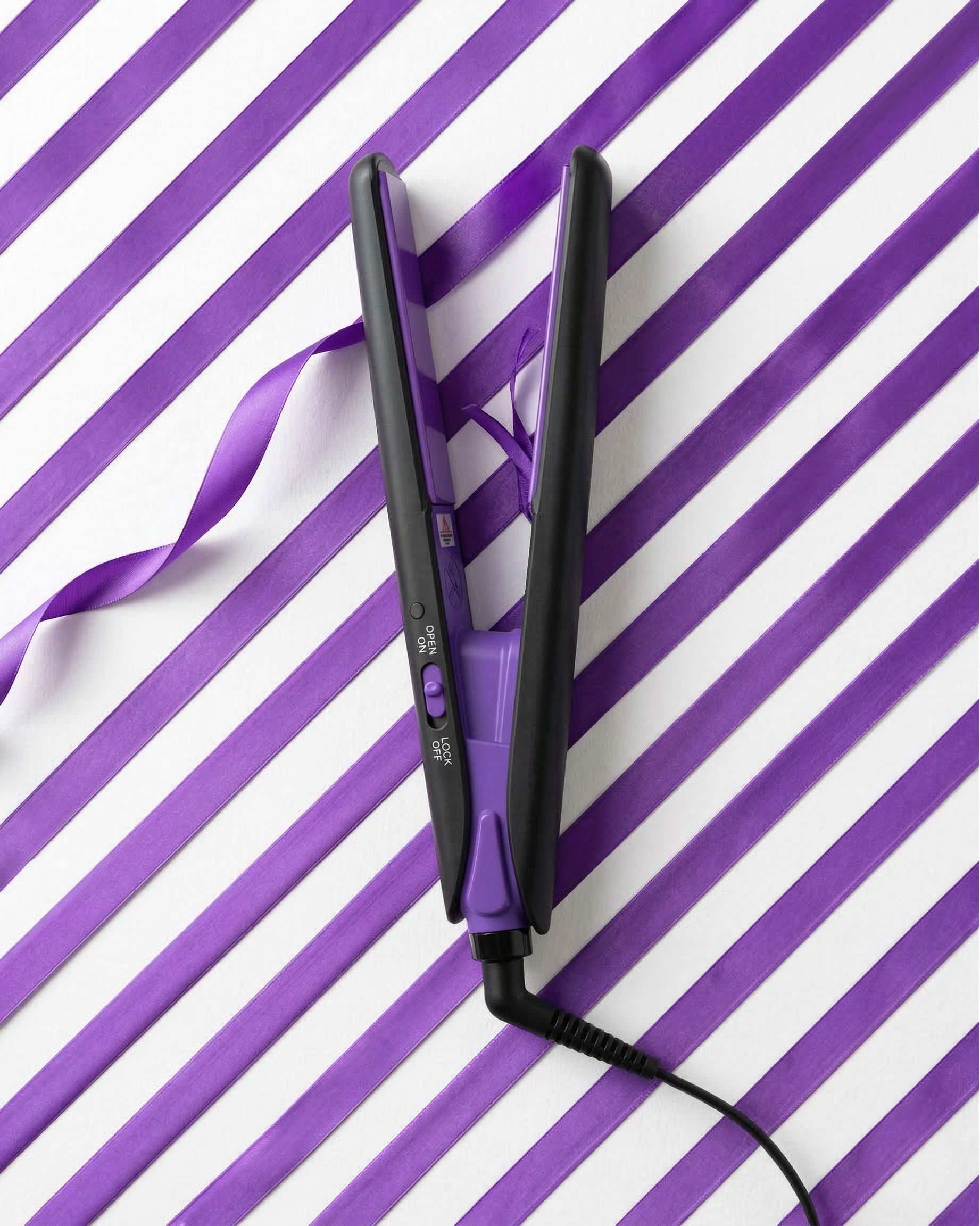 Mini piastra per capelli nera e viola. Piastre aperte, connessione via cavo. Su motivo a strisce viola e bianco.