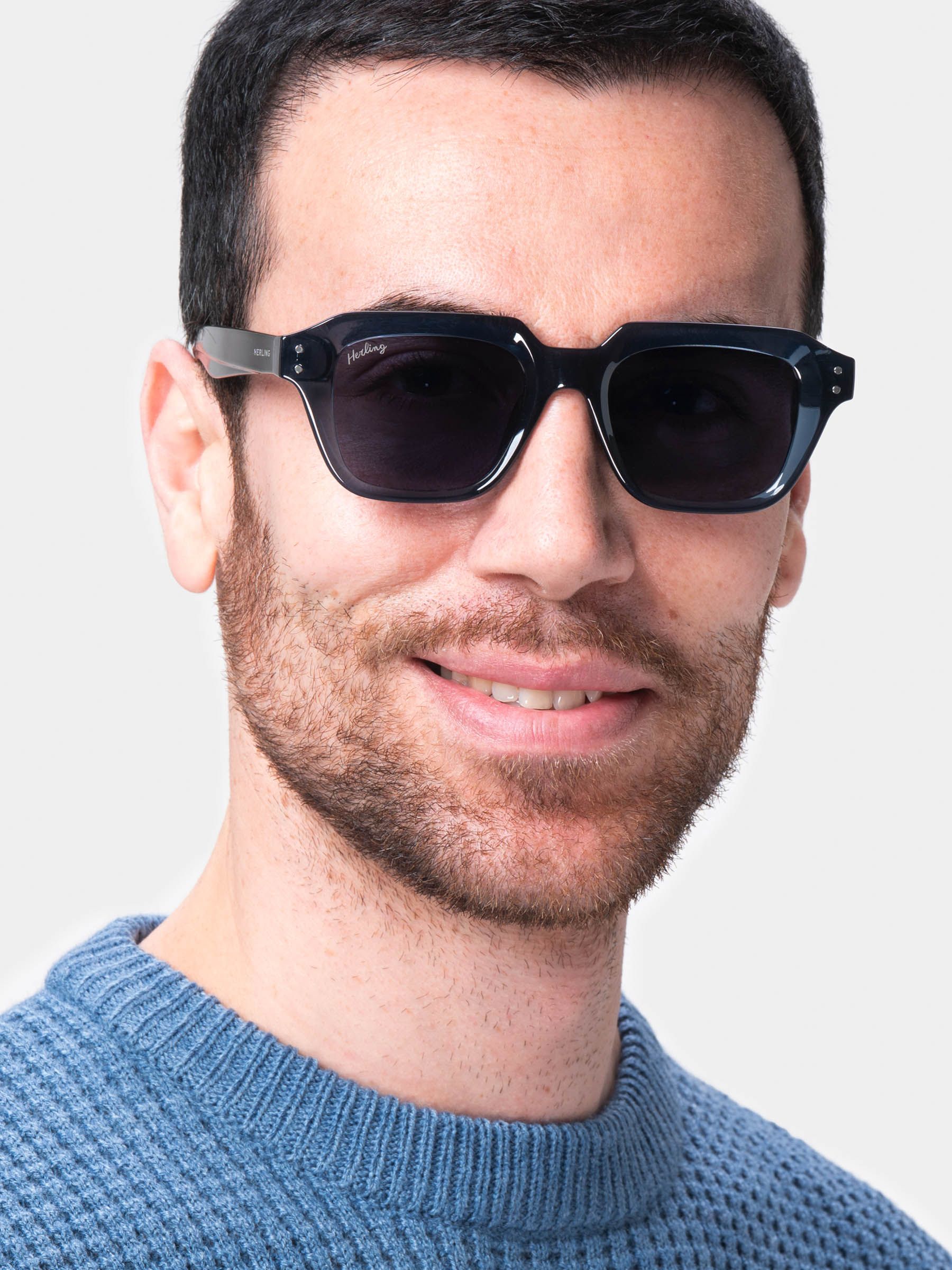 Uomo con occhiali da sole blu scuro. Logo Herling sulla stanghetta. Lenti quadrate. Maglione azzurro.