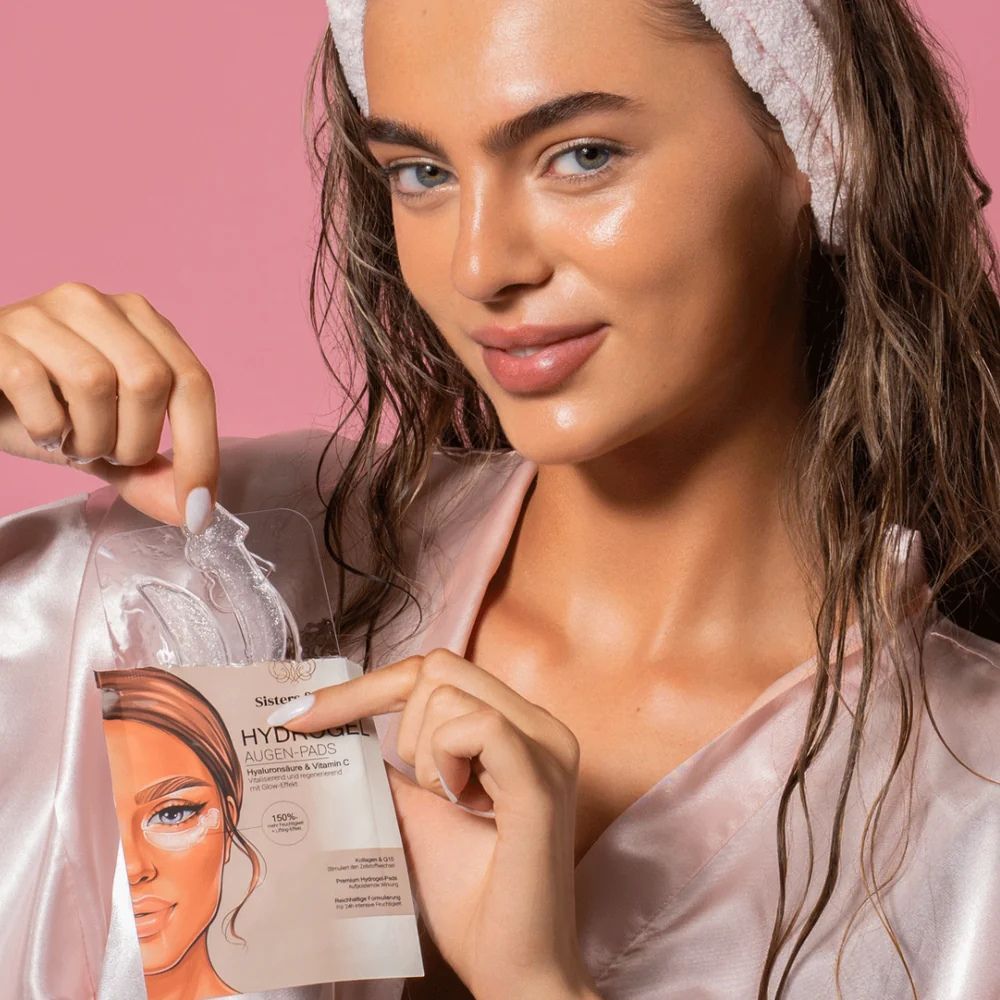 Donna tiene in mano una confezione di cuscinetti idrogel per occhi. Scritta: Sisters & Me, Hydrogel Augen-Pads con acido ialuronico e vitamina C. Cuscinetti visibili.