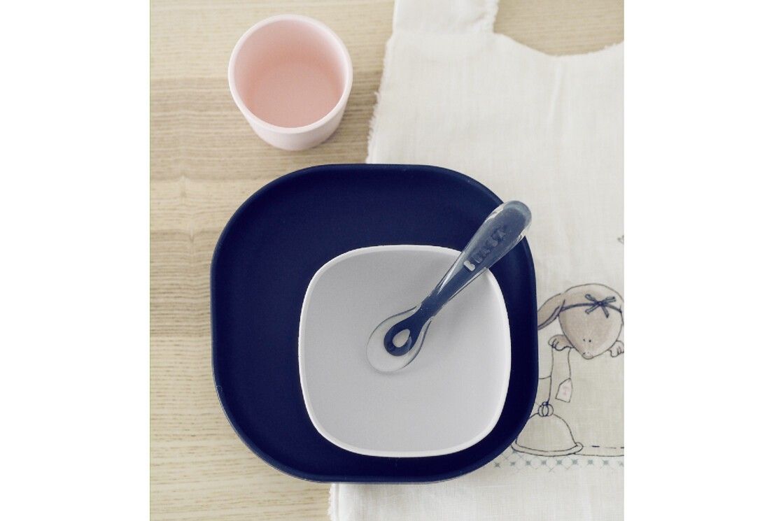 Set di stoviglie BEABA 4 pezzi. Piatto, ciotola, tazza e cucchiaio. Blu, bianco e rosa. Posizionato su tavolo in legno.