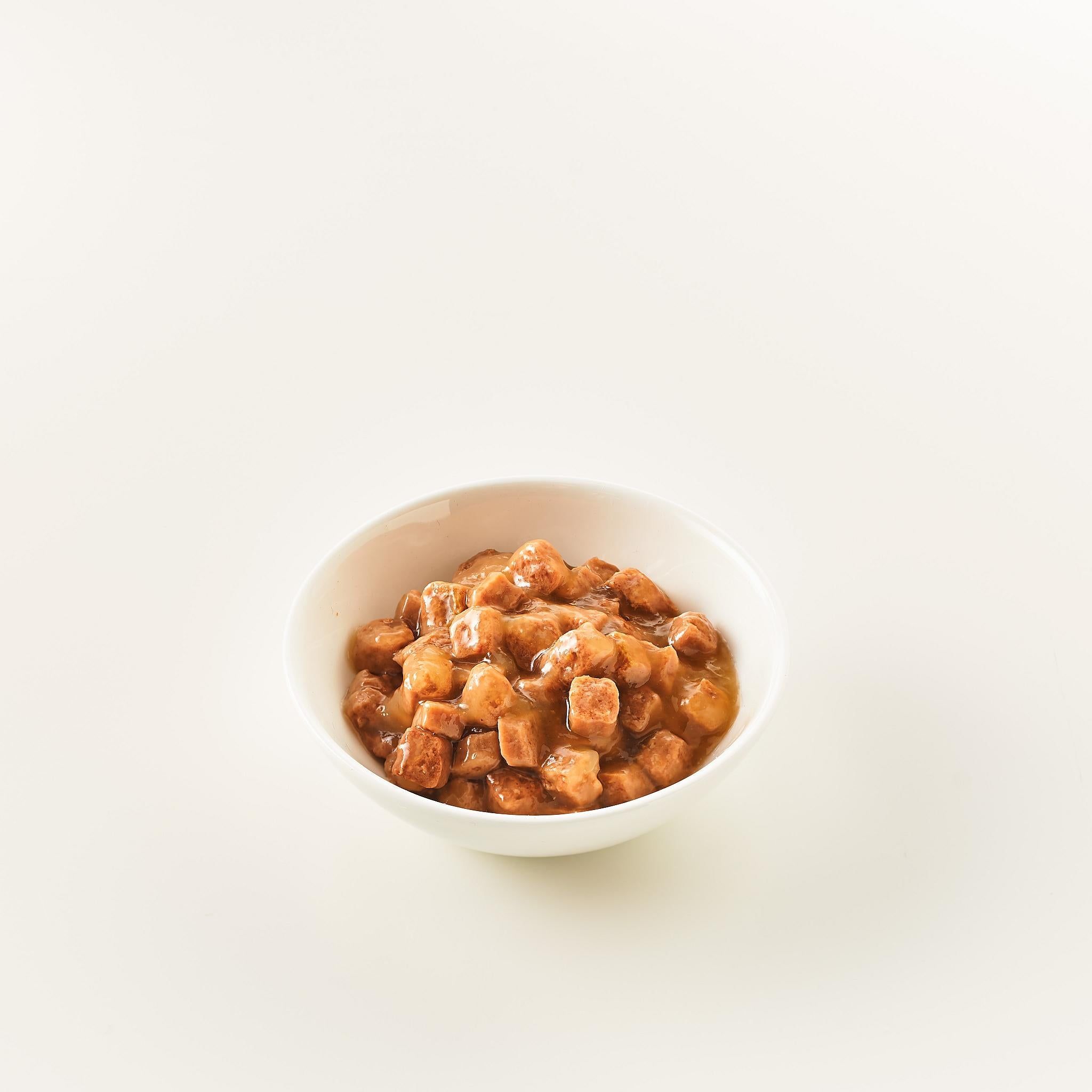 Ciotola bianca con cibo a cubetti marrone. Il cibo è in una salsa marrone chiaro. Su sfondo bianco.
