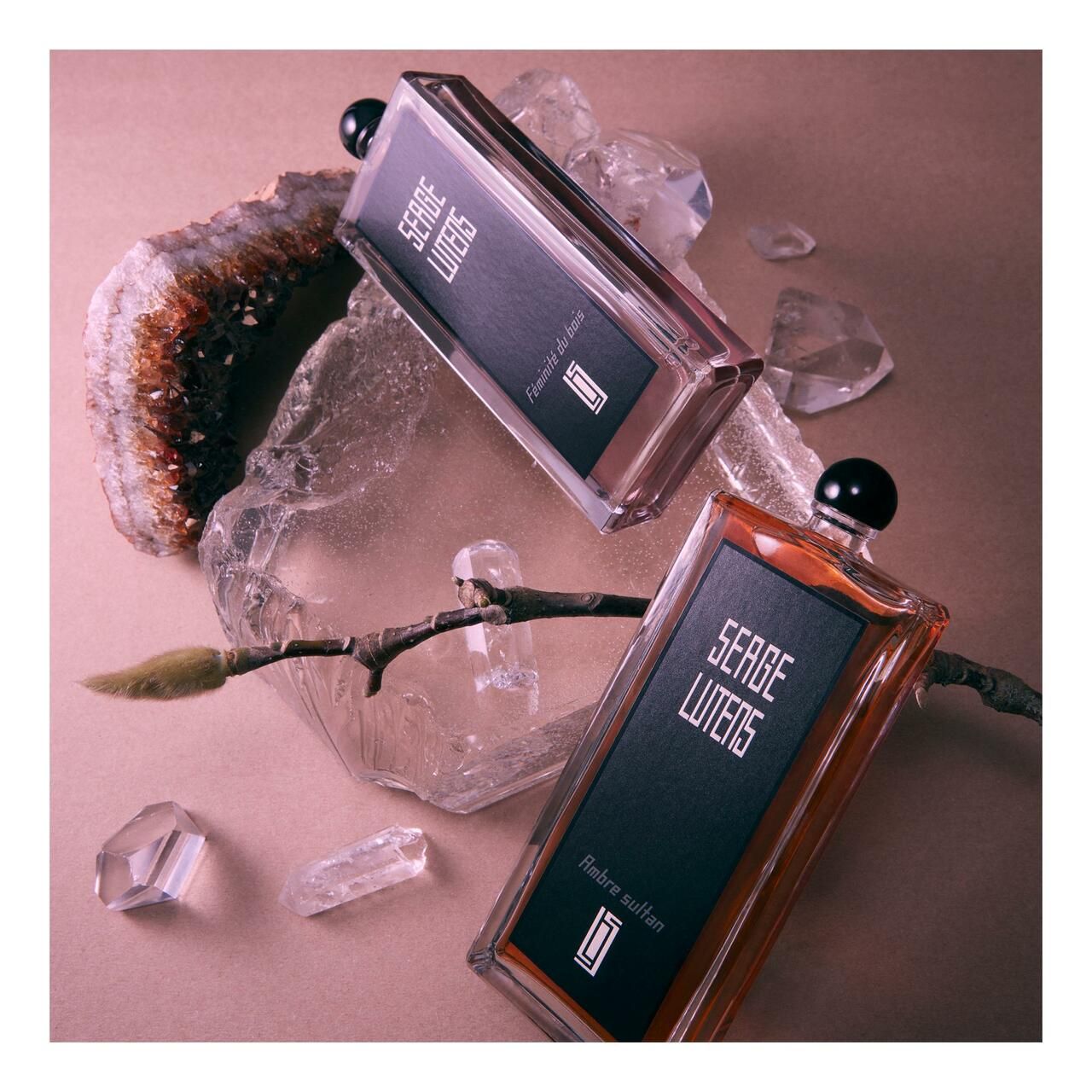 Due flaconi Serge Lutens su una superficie con cristalli e rami. Un flacone è ambrato, l'altro rosa.