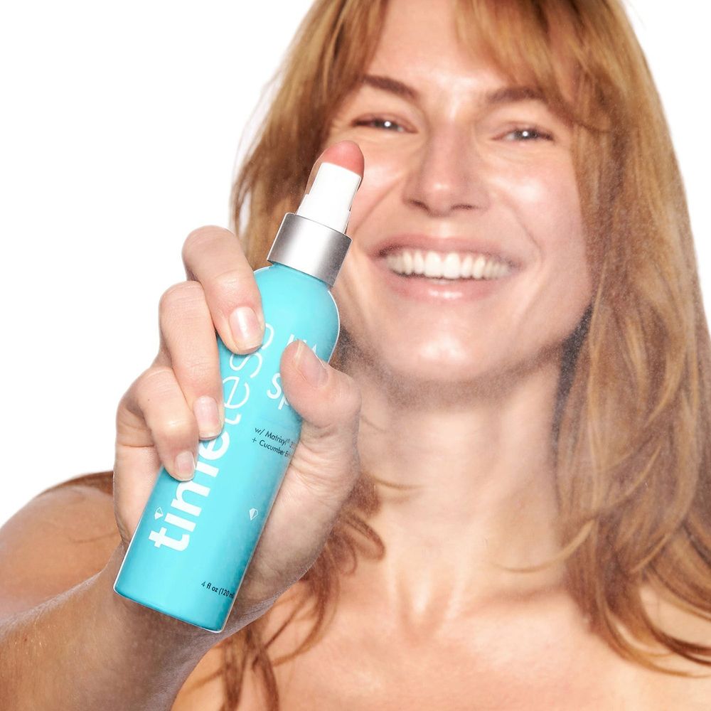 Donna che spruzza con un flacone turchese. Scritte: timeless, HA Spray, Matrixyl 3000 + Cucumber Extract. Nebbia spray visibile.