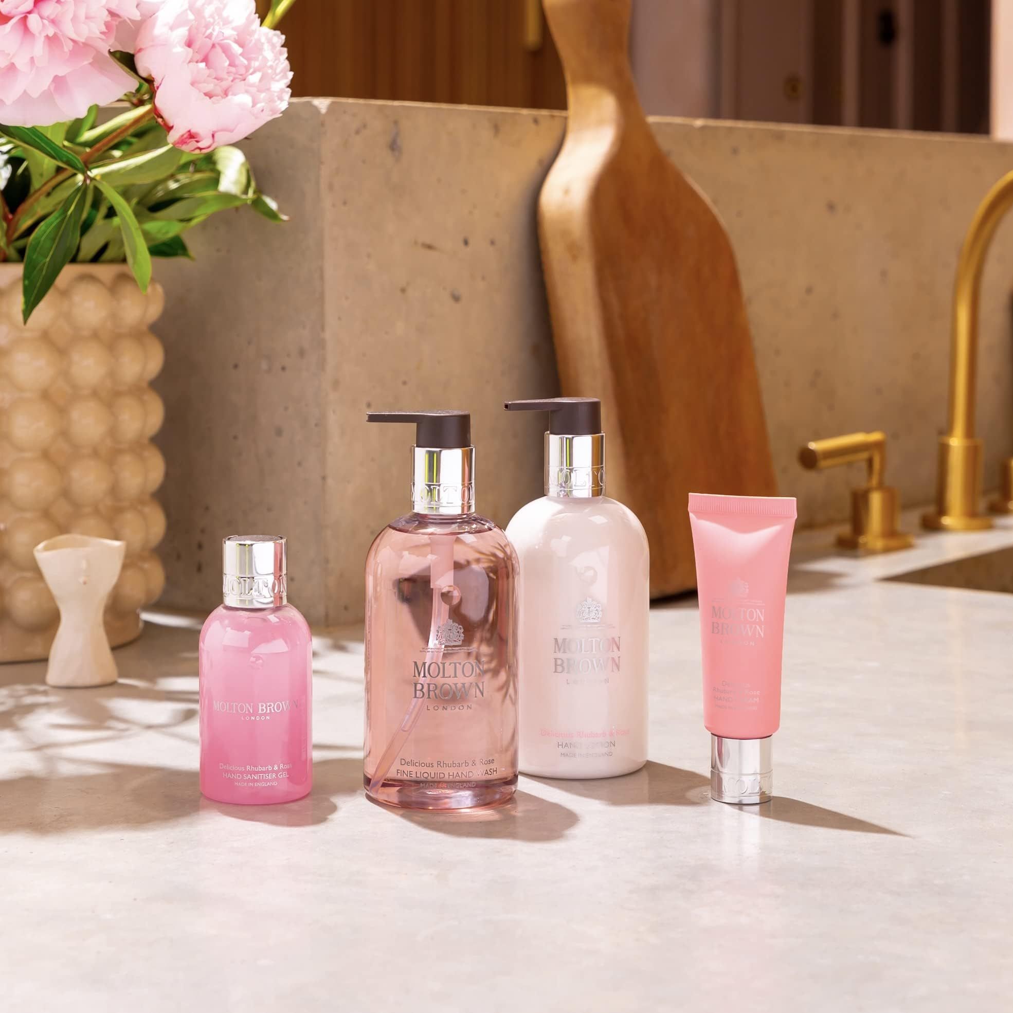 Prodotti rosa su un bancone: crema mani, sapone e profumo. Composizioni floreali sullo sfondo. Rubinetteria dorata.