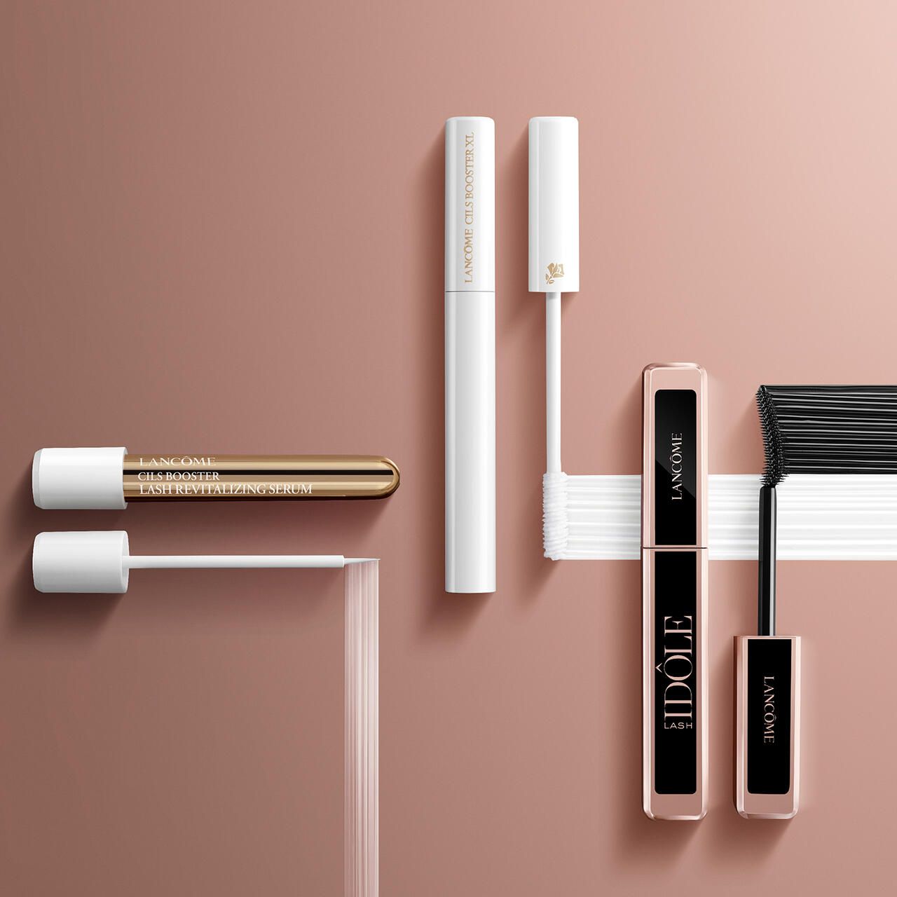 Diversi mascara e primer su sfondo rosa. Custodie dorate e bianche visibili. I prodotti sono disposti.