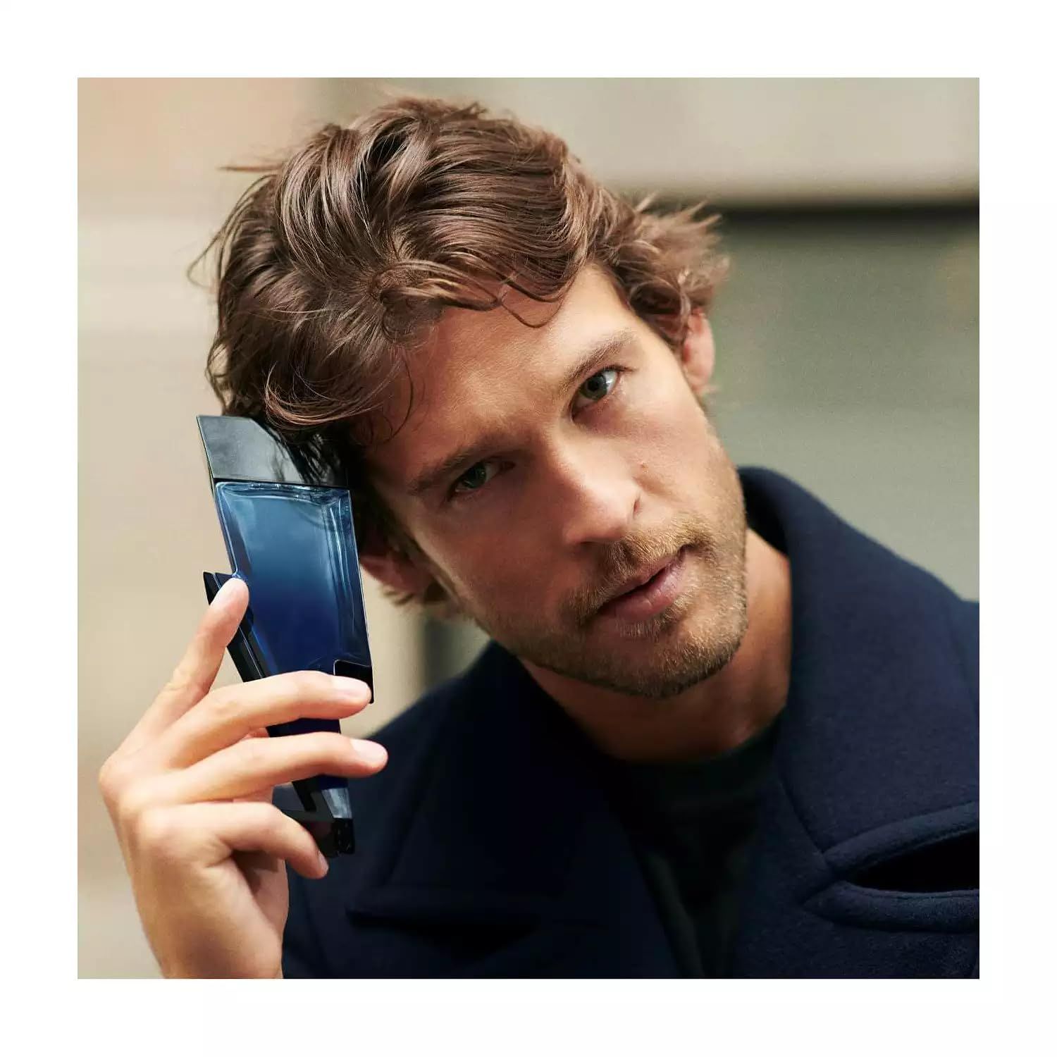 Uomo tiene in mano flacone blu a forma di fulmine. Indossa un cappotto blu e guarda la fotocamera.