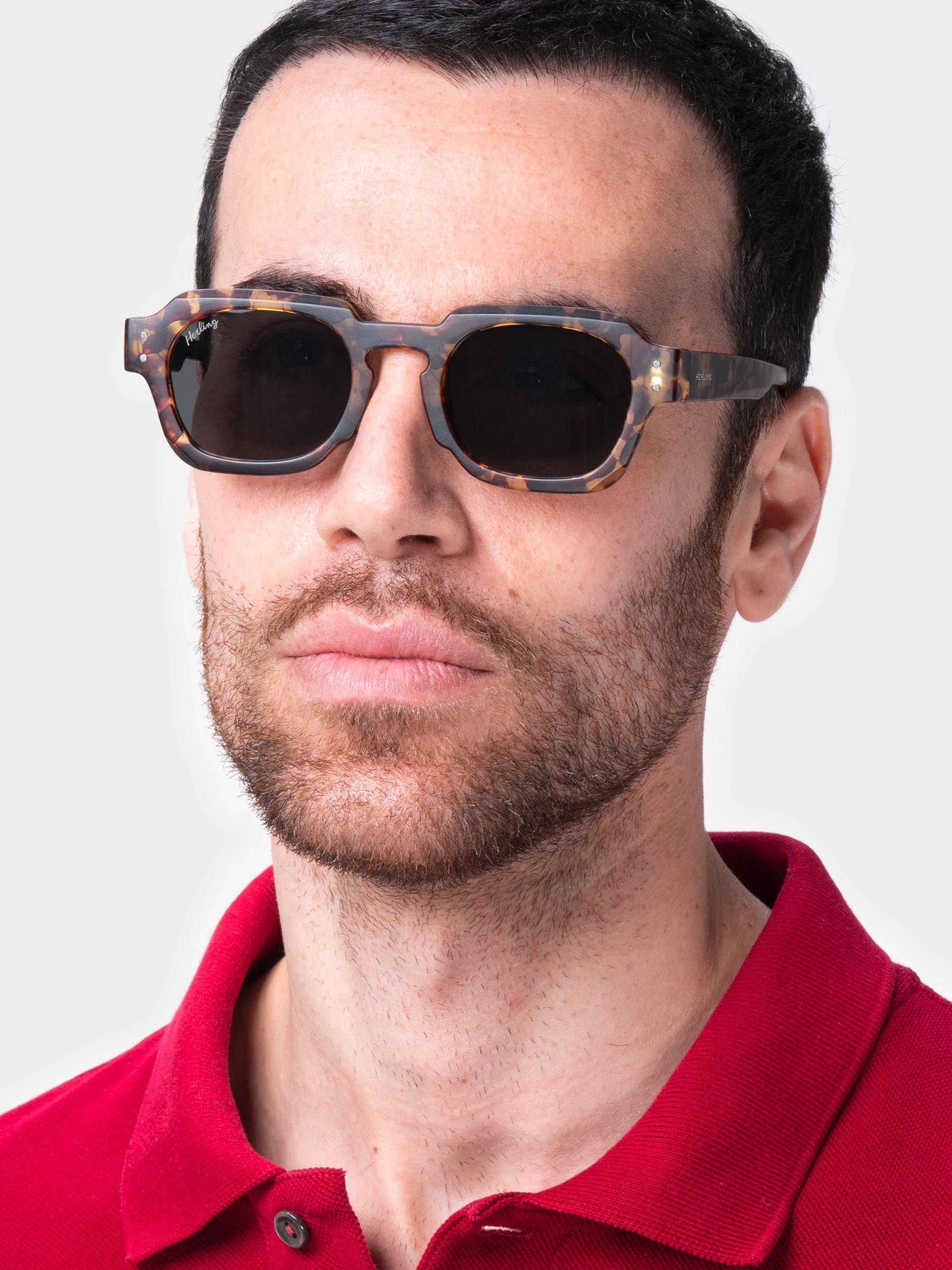Primo piano di un uomo con occhiali da sole. Fantasia tartarugata marrone, lenti scure, forma squadrata. Camicia rossa.