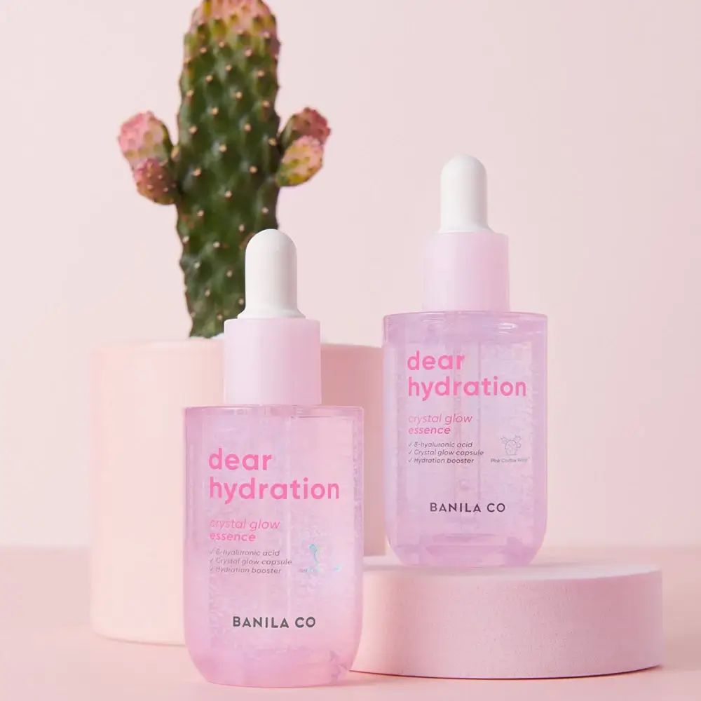 Due flaconi BANILA CO Dear Hydration Crystal Glow Essence. Flaconi rosa con tappi bianchi, cactus sullo sfondo.