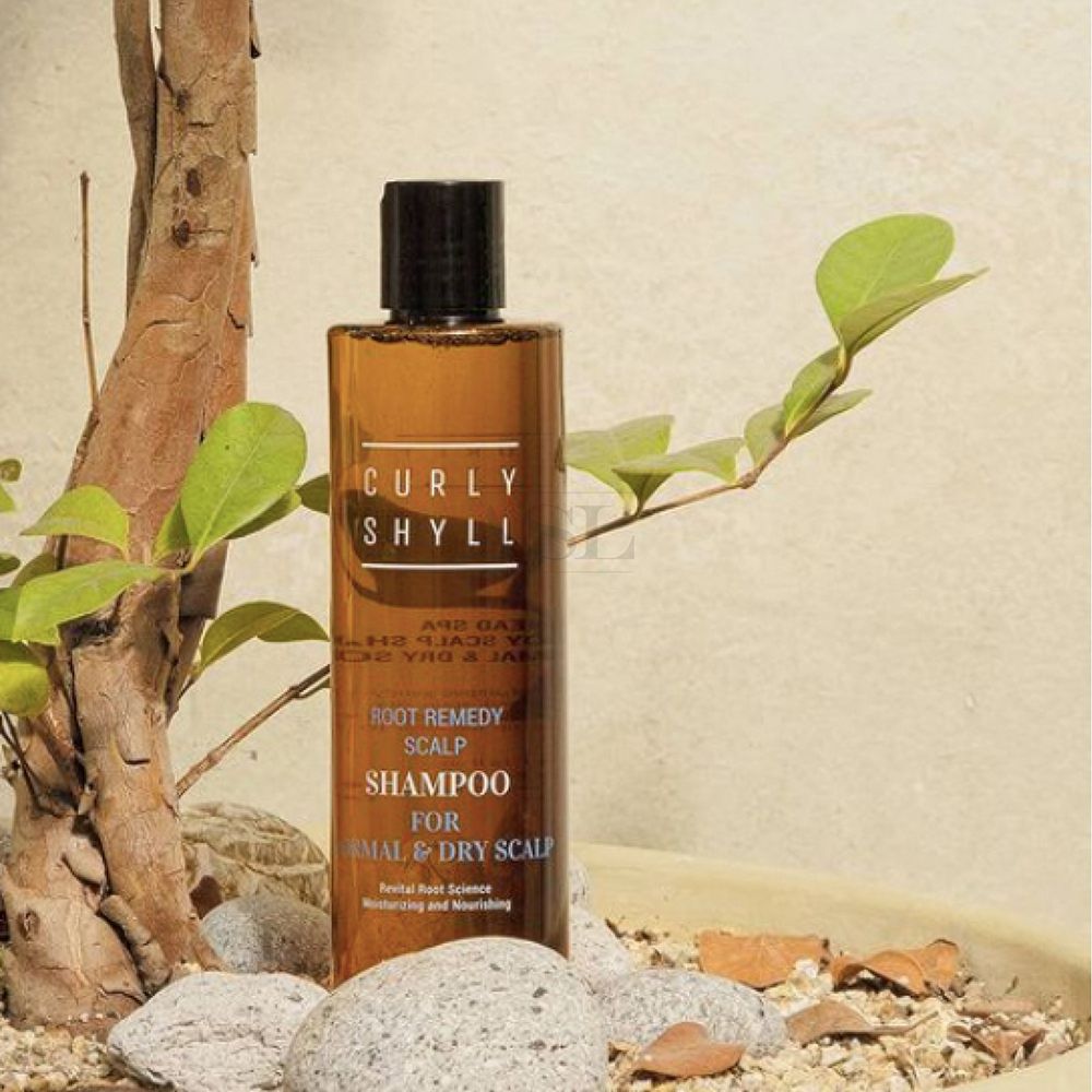 Flacone di shampoo marrone con tappo nero. Scritta: CURLY SHYLL, Root Remedy Scalp Shampoo, per cuoio capelluto normale e secco. Circondato da piante.