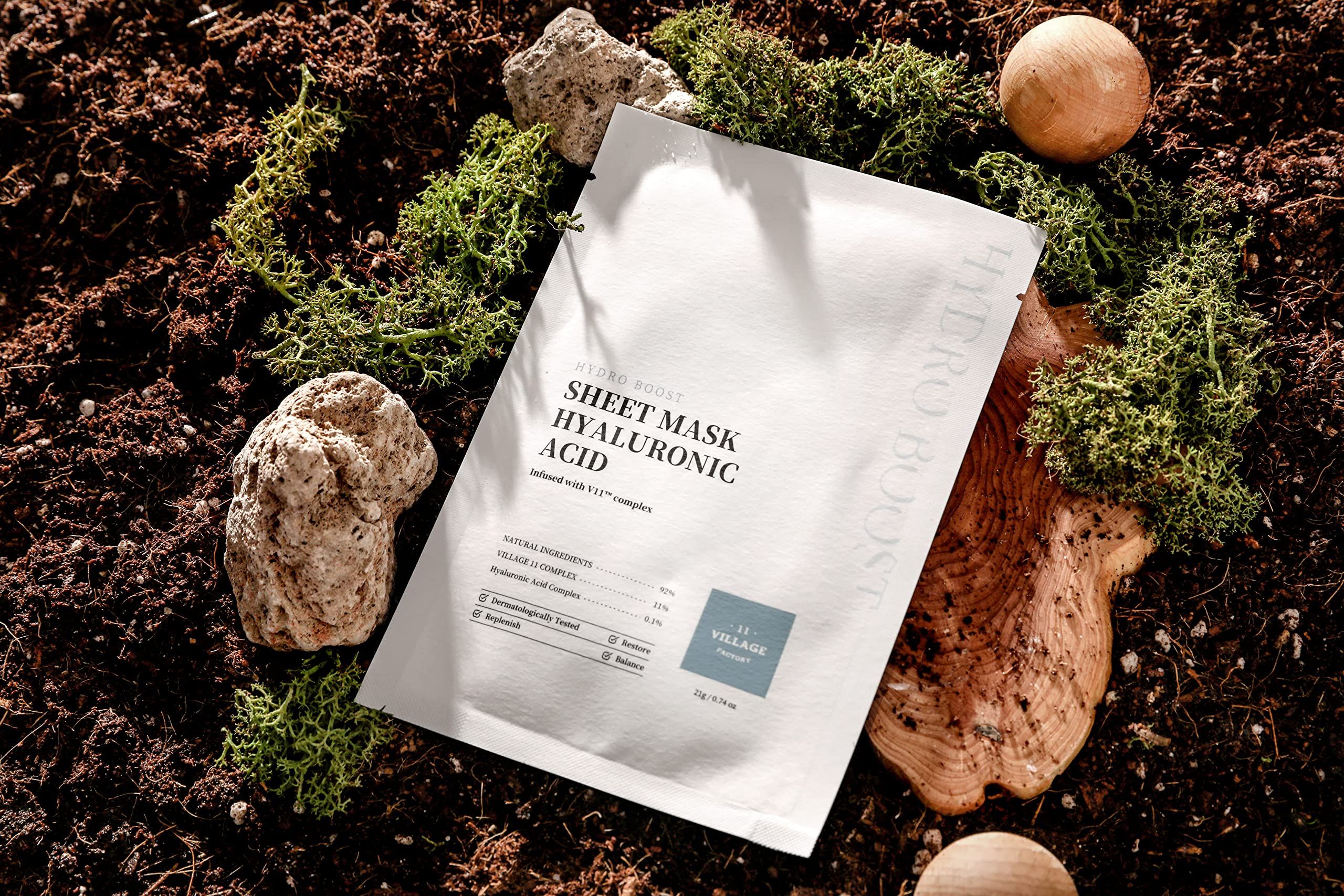 Maschera in tessuto bianca con testo. Scritta: Sheet Mask Hyaluronic Acid. Posizionata su terra con muschio e pietre.