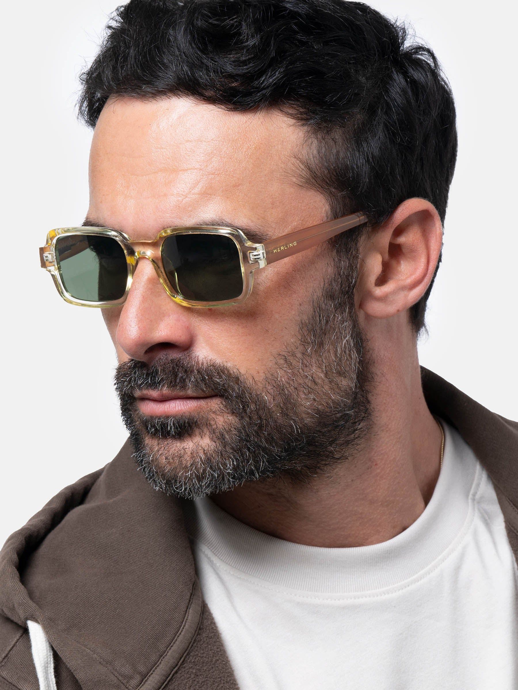 Uomo con occhiali da sole. Gli occhiali hanno una montatura giallo chiaro e lenti verdi. L'uomo indossa una felpa con cappuccio marrone.