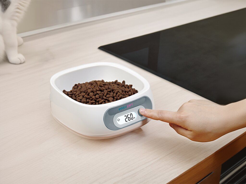 Ciotola bianca con bilancia integrata, riempita con cibo secco. Una mano preme il pulsante della bilancia.