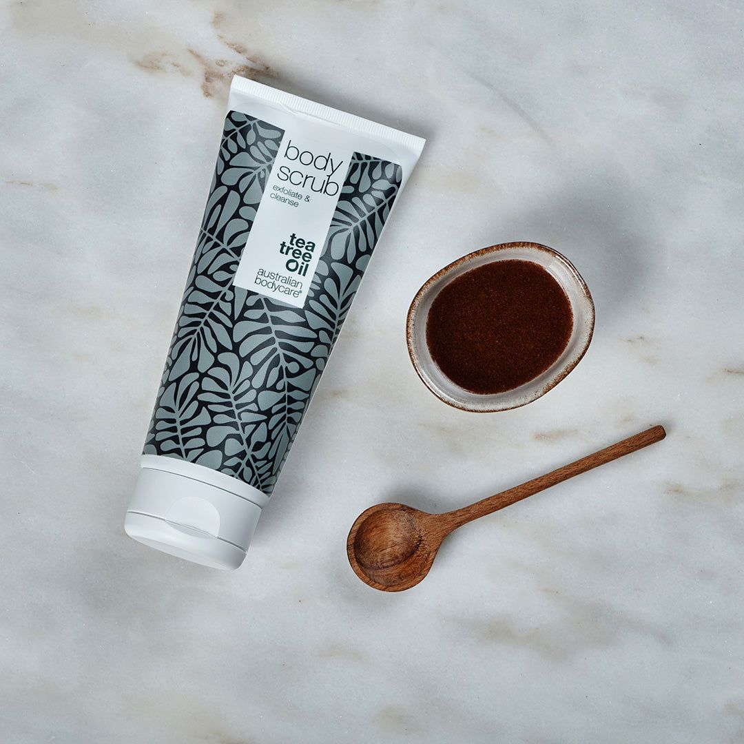 Tubo di Body Scrub, ciotola e cucchiaio. Tubo bianco con motivi a foglie grigie. Testo: Tea Tree Oil, Australian Bodycare. Prodotto marrone.