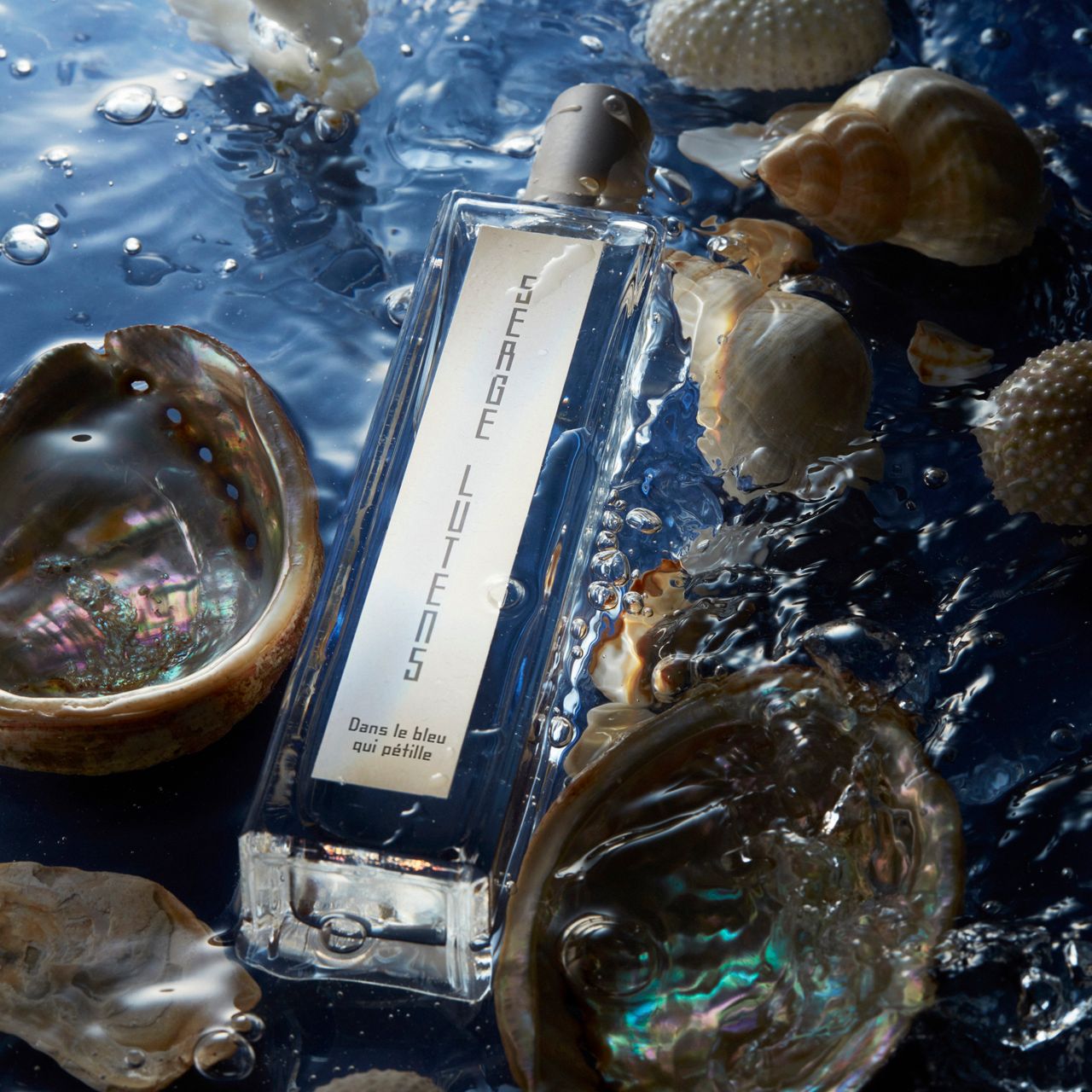Flacone rettangolare in vetro nell'acqua con conchiglie. Scritta: SERGE LUTENS, Dans le bleu qui pétille. Tappo beige.