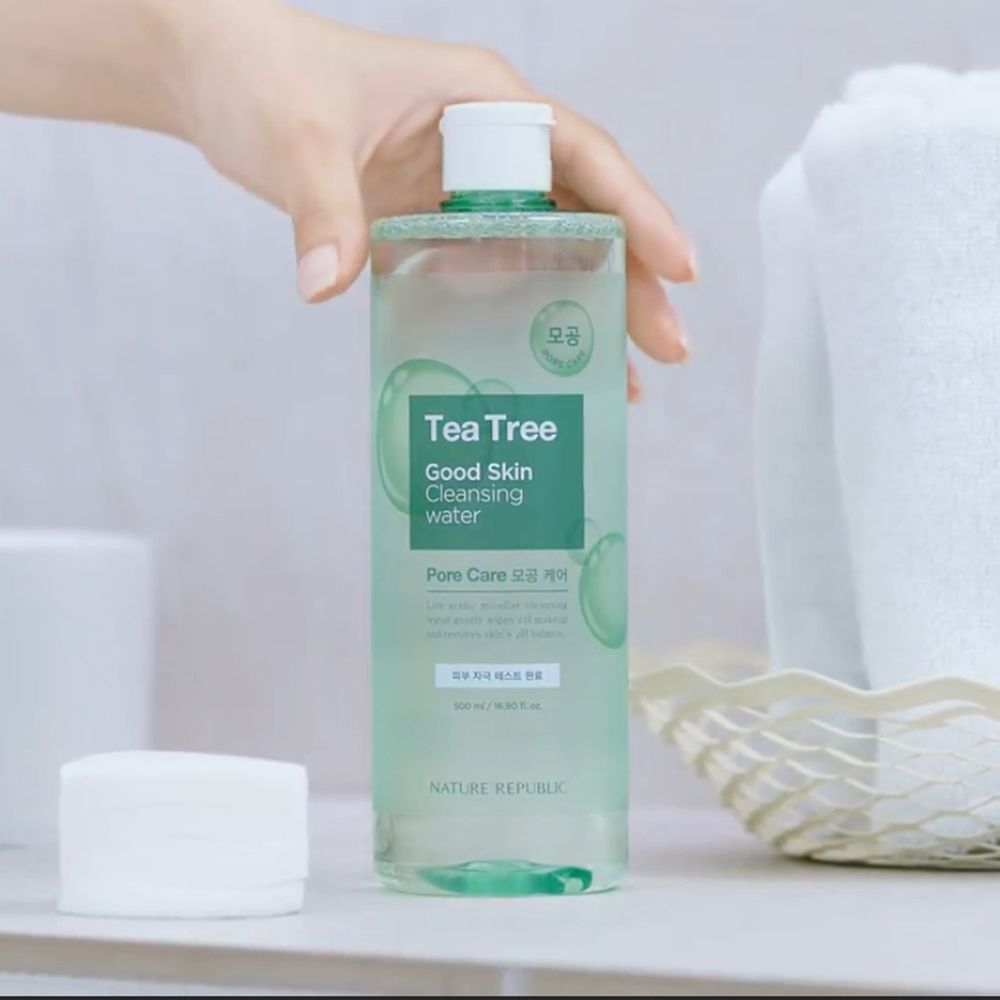Flacone trasparente con etichetta verde. Scritta: Tea Tree Good Skin Cleansing Water. Mano che tiene il flacone. Dischetti di cotone accanto.