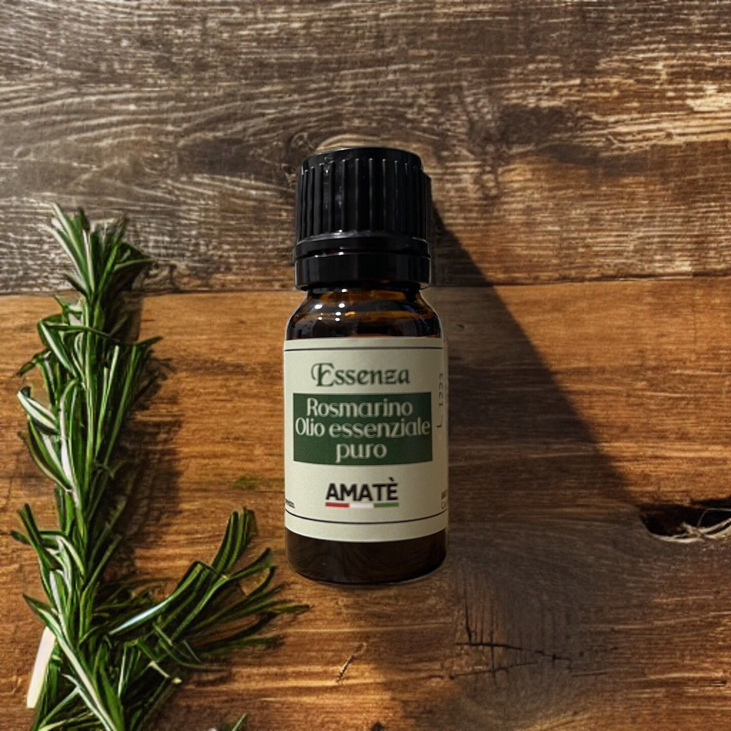 Flacone di vetro marrone con tappo nero, accanto a rametti di rosmarino. Etichetta: Essenza Rosmarino Olio essenziale puro. Marchio: AMATÈ.