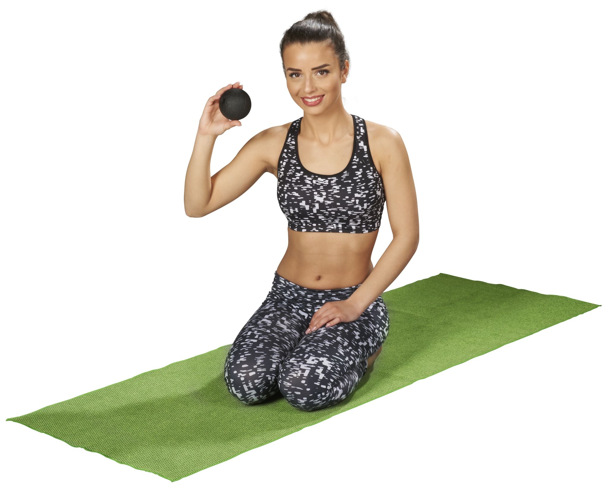 Donna inginocchiata su tappetino verde, tiene in mano una sfera per fascia nera. Abbigliamento sportivo. Sfondo bianco.