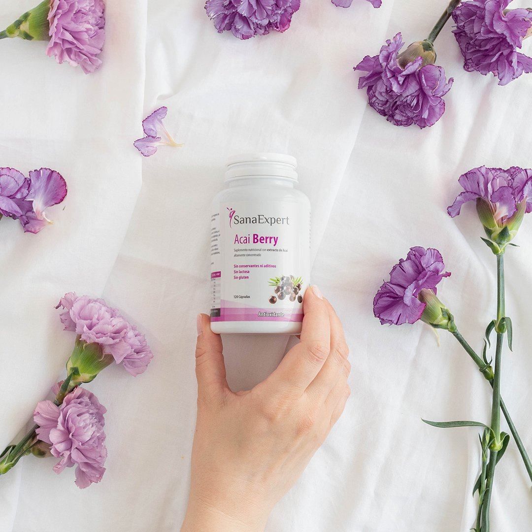 Una mano tiene una bottiglia di Acai Berry, circondata da fiori viola su tessuto bianco. L'etichetta del prodotto è visibile.