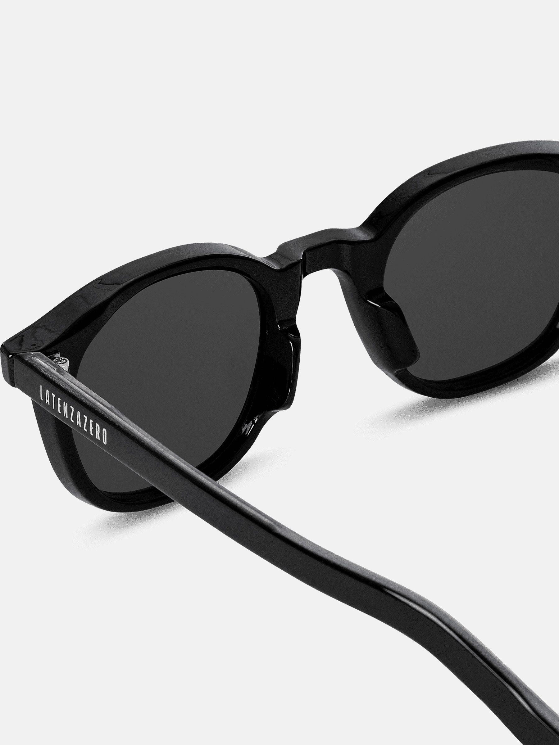 Primo piano degli occhiali da sole neri. Lenti rotonde, montatura e aste nere. Marchio sull'asta.