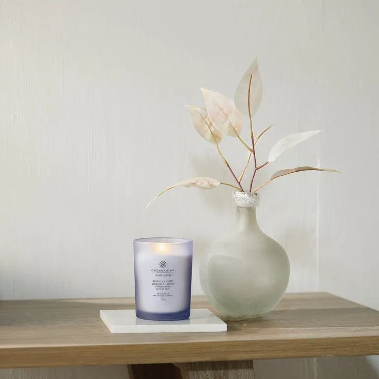Candela blu su sottobicchiere bianco. Vaso con ramo accanto. Scritta: Chesapeake Bay, serenity + calm.