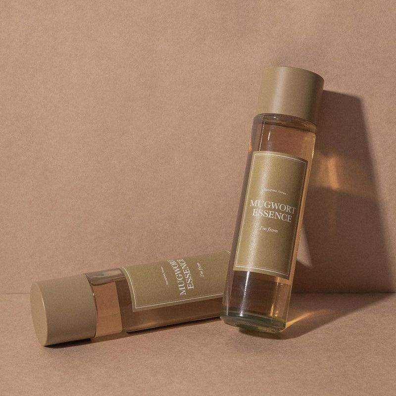 Due bottiglie con etichette verdi. Scritta: MUGWORT ESSENCE, I'M FROM. Sfondo beige.