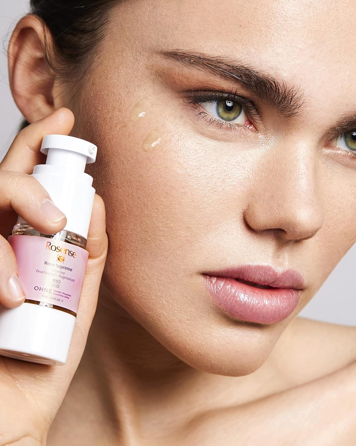 Donna tiene Rosense Supreme Augenserum. Dispenser bianco, etichetta rosa, testo. Gocce sul viso. Sfondo: grigio.