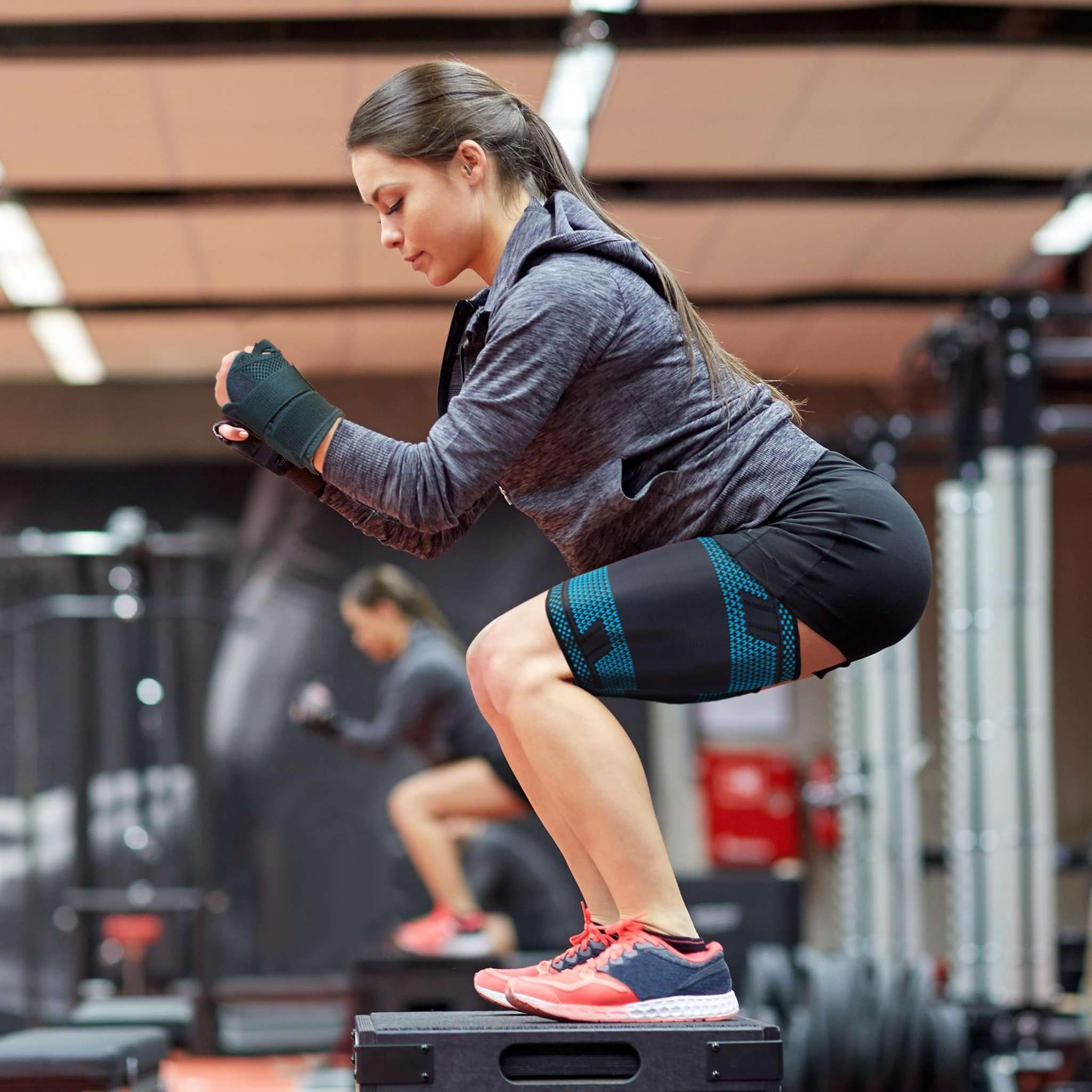 Donna fa squat in palestra. Indossa fasce per coscia AGILE NOW. Nero-turchese. Tappetino ginnico.